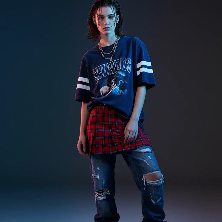 Looking for a trendy oversized women’s graphic T-shirt? A stylish teenager wears a navy blue graphic tee with white accents, paired with a red plaid mini skirt and distressed baggy jeans, perfect for streetwear and casual fashion looks.